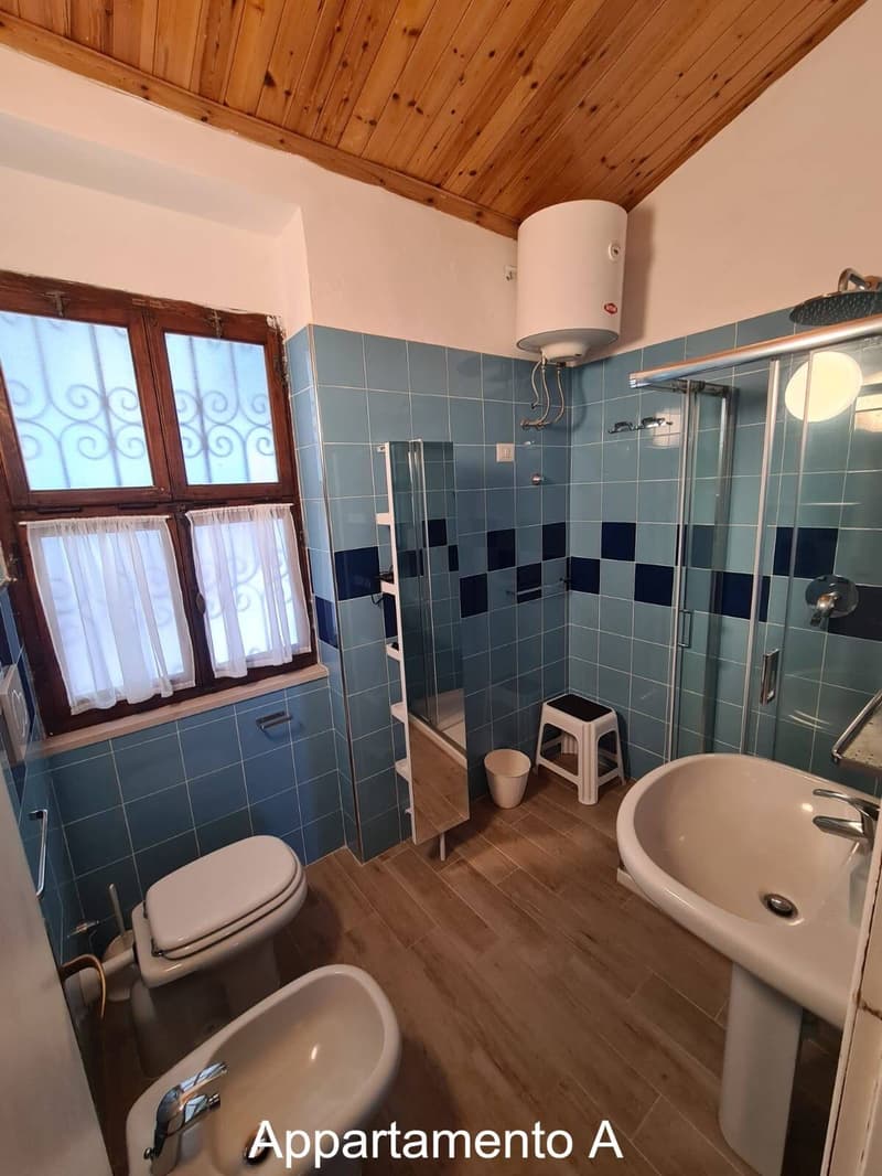 Tiled blue and white wall and floor, window with white curtains, modern shower cubicle, toilet, bidet, sink with a faucet, white storage box, metal shower head.