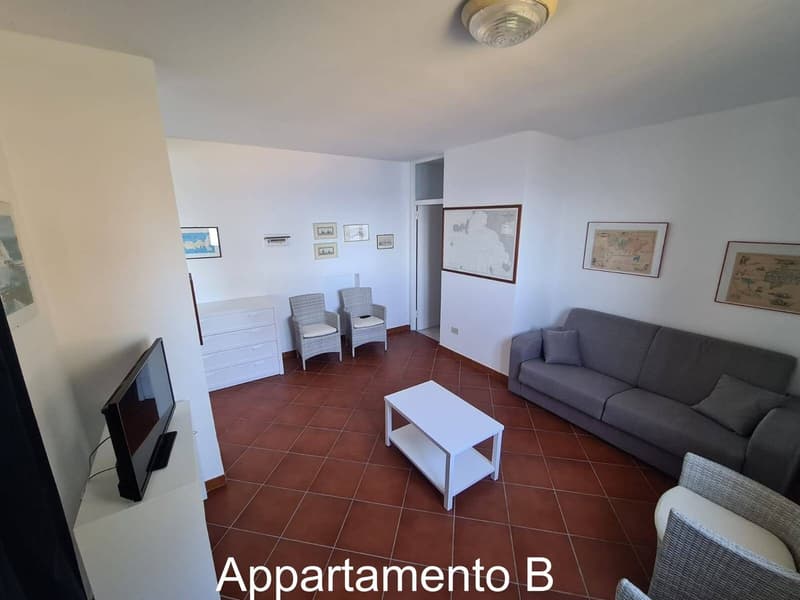 Living room with tiled floor, white walls, TV, coffee table, couch, chairs, white dresser, framed pictures