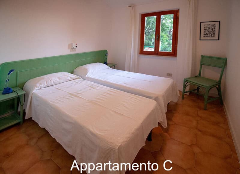 2 single beds, white sheets, green headboards, night stands with lamps, green chair, picture frame, window with curtains, tiled floor