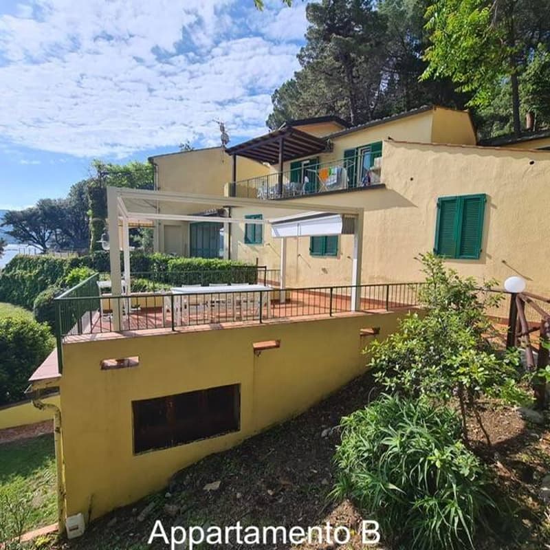 A two-story yellow building with a raised terrace with a canopy, balconies, greenery, sea view, and surrounded by trees