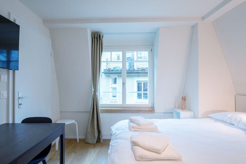 Spacious bedroom with large windows, white walls, hardwood floor, bed, desk, and TV