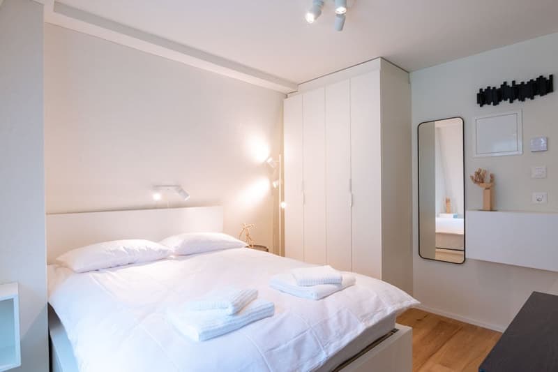 Spacious bedroom with white walls, hardwood floor, large wardrobe, bedside table, and a mirror on the wall