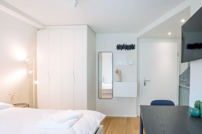 Spacious bedroom with white walls, large wardrobe, mirror, and wooden flooring. The room also has a balcony or terrace visible.
