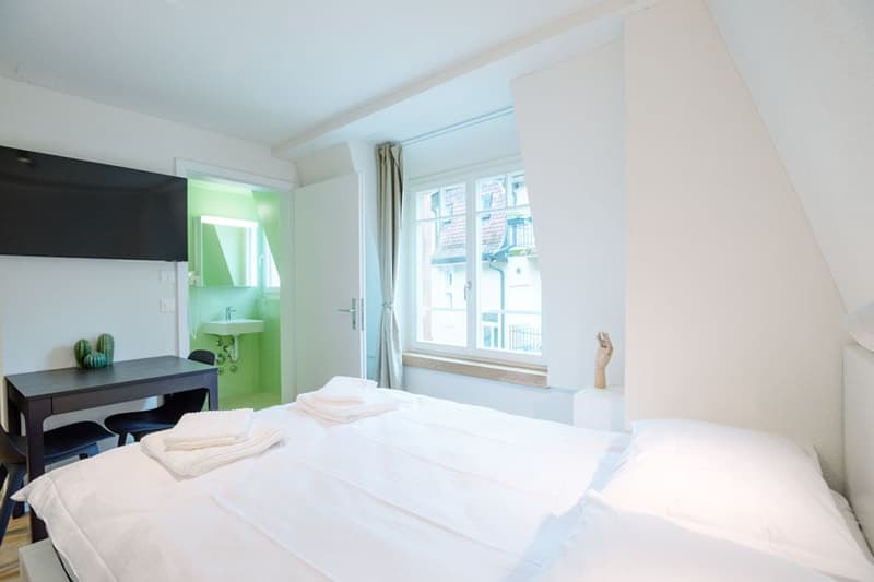 Bright bedroom with large window, white walls, dark furniture, and an en-suite bathroom with green accents