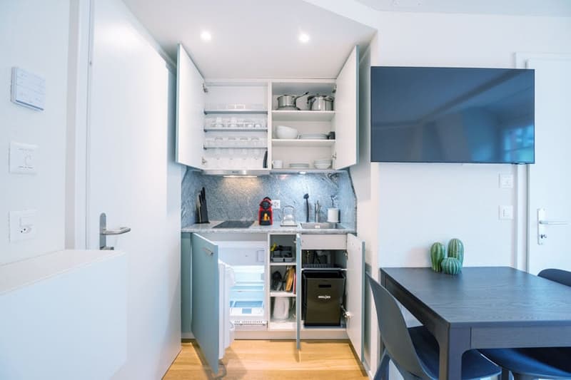 Compact kitchen with white cabinets, open shelving, gray backsplash, built-in dishwasher, and washing machine
