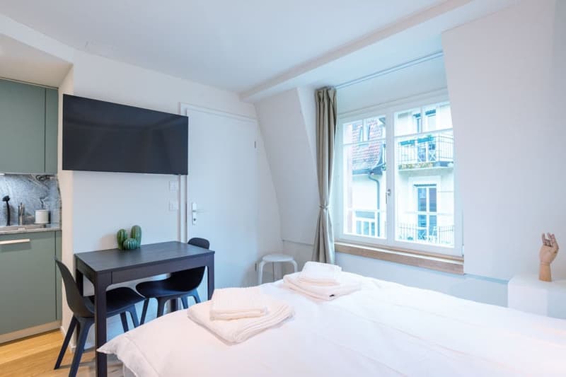 Bright and modern studio apartment with large windows, white walls, hardwood floors, a kitchenette with dark cabinets, a dining table, and a bed with white bedding. The apartment also has a balcony or terrace visible.