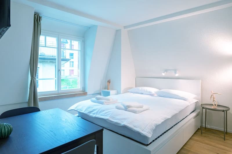 Spacious bedroom with large windows, white walls, hardwood floor, king-size bed, bedside table, and a balcony or terrace visible.