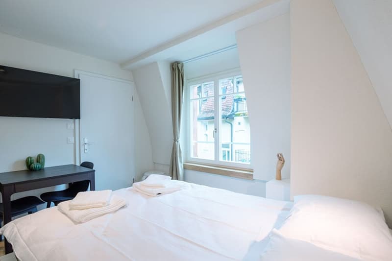 Spacious bedroom with large window, white walls, dark wooden desk, and white bedding