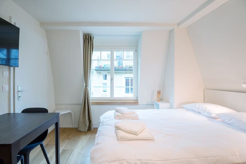 Spacious bedroom with large windows, white walls, hardwood floor, bed with white bedding, desk, and curtains