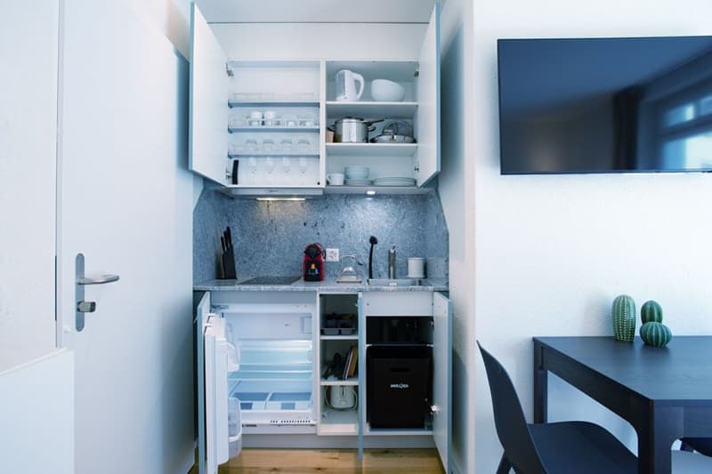Compact kitchenette with open shelving, refrigerator, and dishwasher. The space features a minimalist design with white walls and cabinets.
