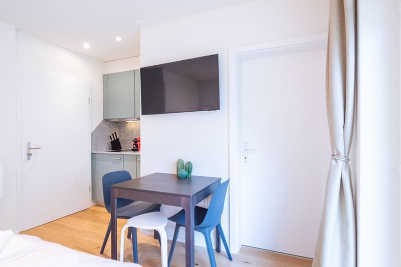 Compact studio apartment with kitchenette, dining table, and mounted TV. The space features hardwood floors, white walls, and modern furnishings including blue chairs and a small kitchen with a dishwasher.