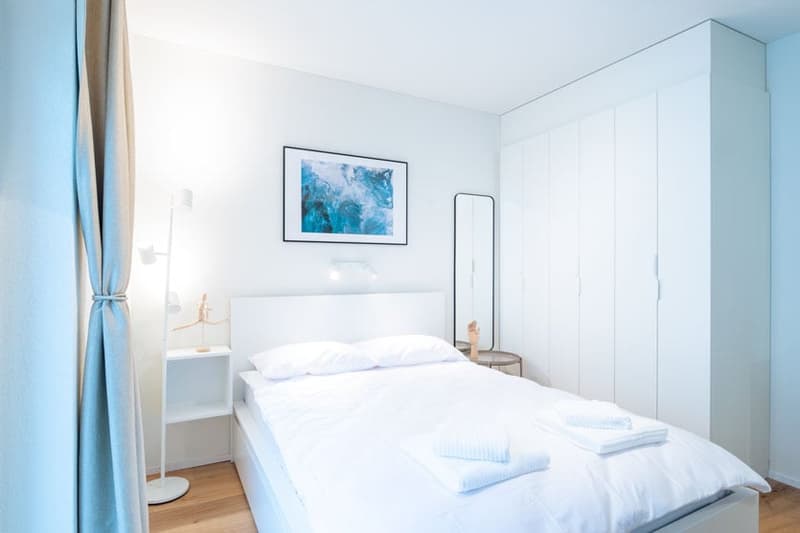 Bright and airy bedroom with white walls, hardwood floors, a large bed with white bedding, a framed artwork on the wall, and a curtained window or balcony door
