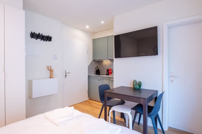 Compact studio apartment with kitchenette, dining table, and TV mounted on the wall. The room has a minimalist and modern design with white walls, hardwood floors, and built-in storage and appliances.