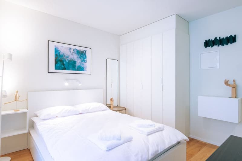 Spacious bedroom with white walls, large bed with white bedding, wooden nightstands, built-in wardrobe, and a framed artwork on the wall