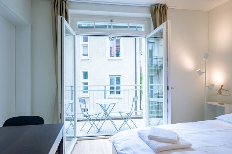 Spacious bedroom with large windows and a balcony, white walls, hardwood floor, bed, table, and chairs