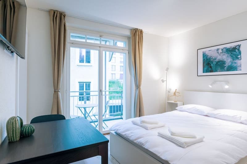 Bright and airy bedroom with large windows, balcony access, white walls, hardwood floor, and modern furniture including a desk and a cactus decoration.