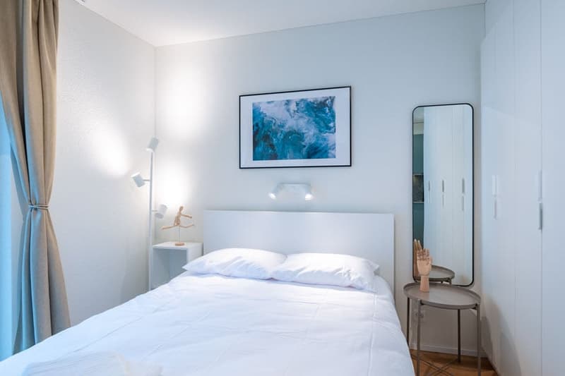 Spacious bedroom with white walls, large framed artwork above the bed, bedside table, and a full-length mirror on the wall.