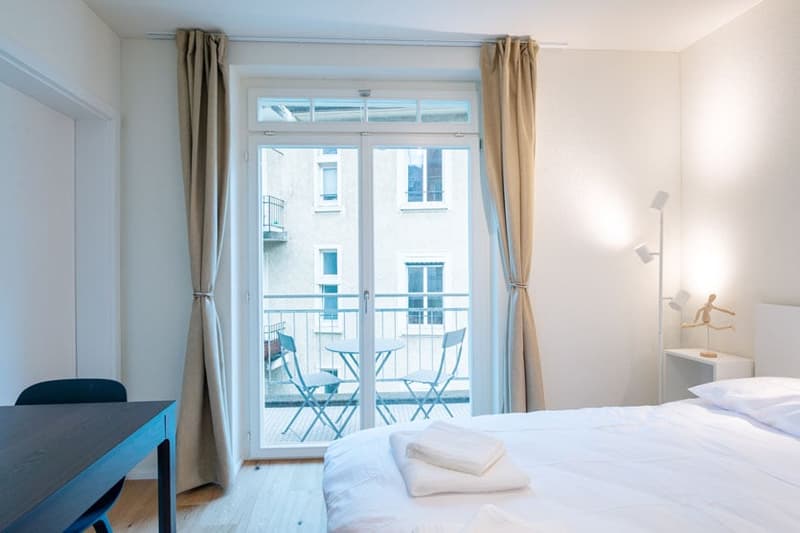 Bright and airy bedroom with large windows, balcony access, wooden floor, and minimalist furniture including a desk and chairs