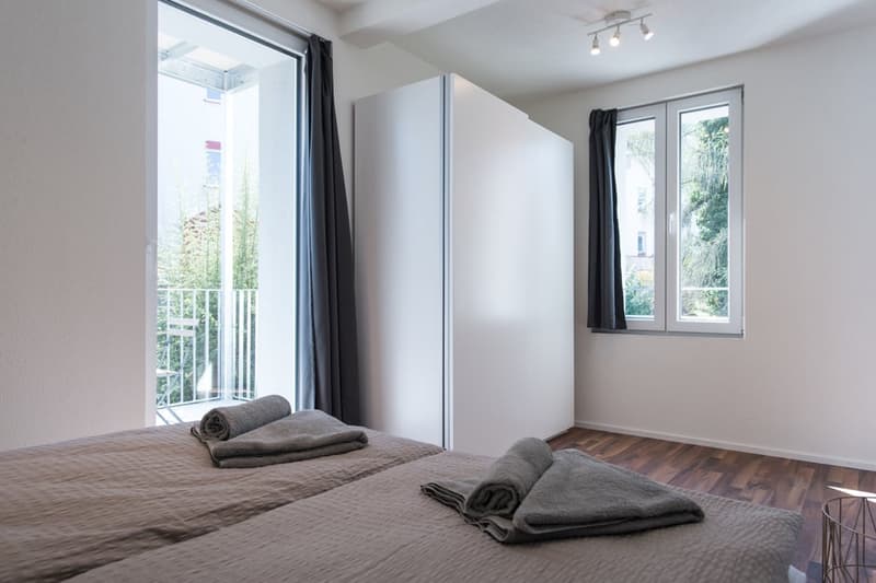 Spacious bedroom with large windows, hardwood flooring, built-in closet, and a balcony or terrace overlooking the greenery outside