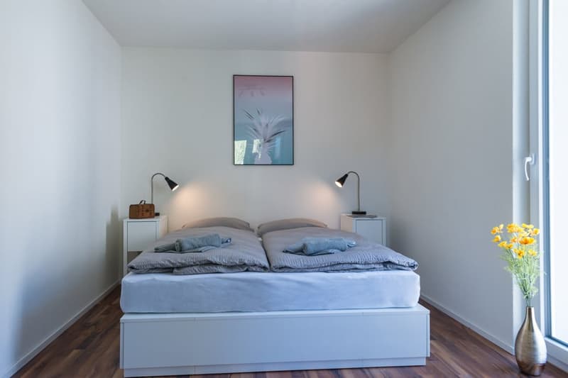 The image shows a modern and minimalist bedroom with a large bed, two bedside tables with lamps, and a framed artwork on the wall. The room has white walls, hardwood flooring, and a large window that provides natural light.