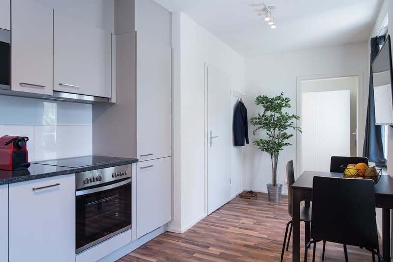 Modern kitchen with white cabinets, black countertop, built-in oven, and dishwasher. Open to a dining area with a wooden table and chairs.