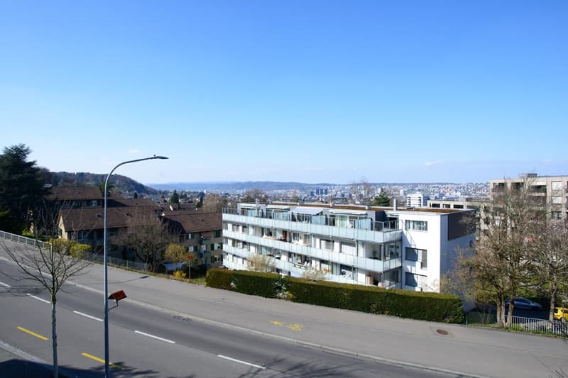 Multi-story apartment building with balconies, surrounded by trees and overlooking a city skyline in the distance.