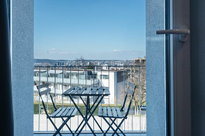 The image shows a balcony or terrace with a view of the city skyline in the distance. There are two black metal folding chairs on the balcony, and the background shows a cityscape with buildings and a clear blue sky.