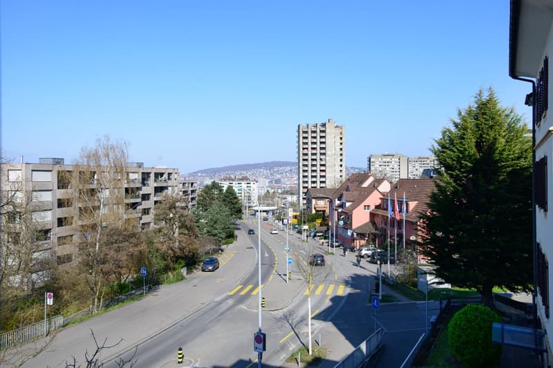 The image shows a cityscape with a wide street, multi-story residential and commercial buildings, trees, and a clear blue sky. The buildings have a mix of architectural styles, and the street appears to be well-maintained with road markings and traffic si
