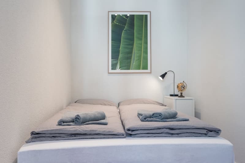 The image shows a simple and minimalist bedroom setup. The room has white walls and a framed artwork featuring green banana leaves. The bed is made with gray bedding, including towels and a comforter. A small side table with a lamp is placed next to the b