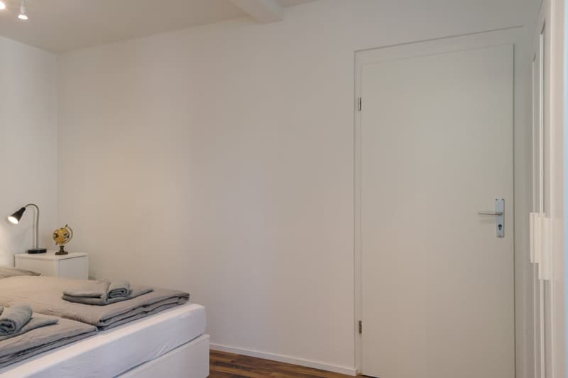The image shows a simple, minimalist bedroom with white walls, a bed with bedding, and a lamp on a side table. There is a door visible on the right side of the room.