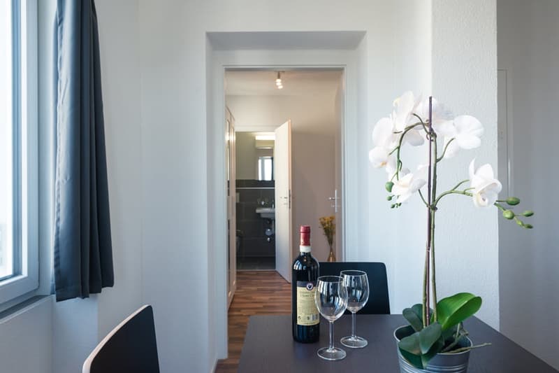 Spacious living room with hardwood floors, white walls, large windows, a dining table with wine glasses and a bottle of wine, and a potted orchid plant