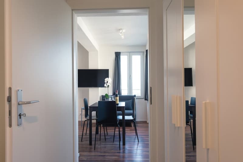 Open concept living room and dining area, hardwood floors, large windows, TV mounted on the wall, dining table with chairs, kitchen appliances visible in the background