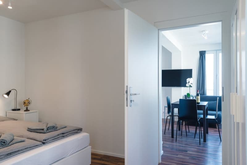 The image shows a modern and minimalist apartment interior. The room in the foreground appears to be a bedroom, with a bed and bedding visible. The doorway leads to an open-plan living and dining area, which can be seen in the background. The apartment ha