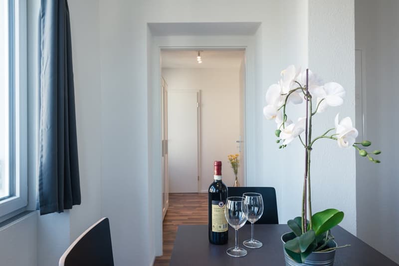 Bright and modern living room with hardwood floors, white walls, large windows, a dining table with wine glasses and a bottle of wine, and a decorative orchid plant.