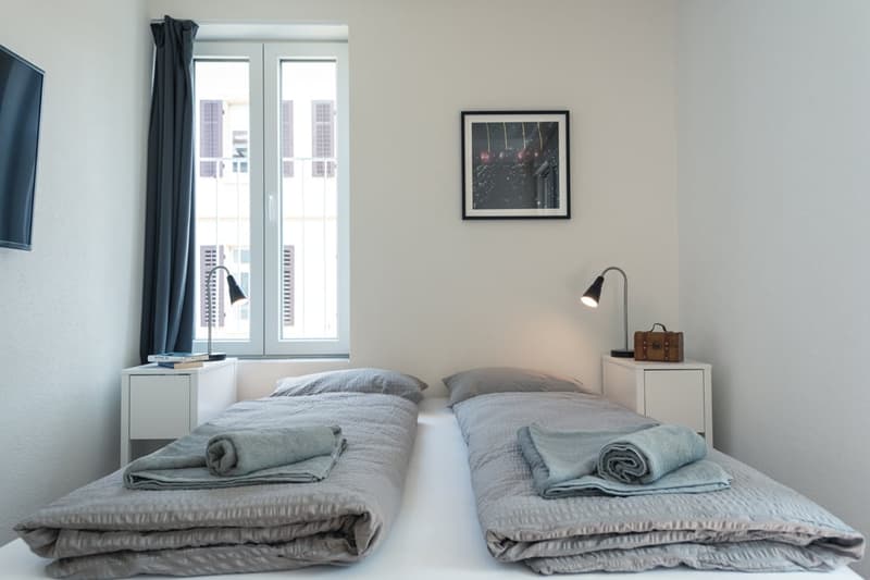 Minimalist bedroom with two twin beds, gray bedding, white walls, large windows, framed artwork, and a bedside lamp