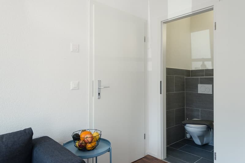 White walls, hardwood floor, sofa, side table with fruit basket, doorway leading to a bathroom with dark tile flooring