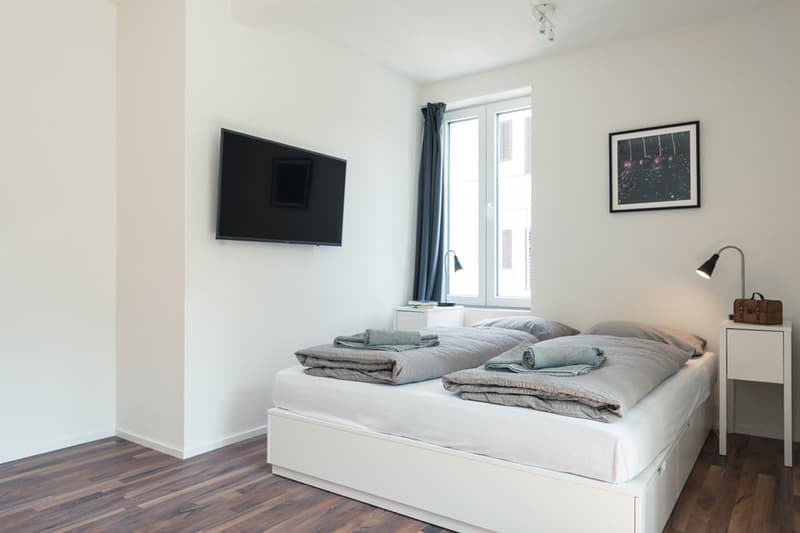 Spacious bedroom with white walls, hardwood flooring, a large bed with grey bedding, a flat-screen TV mounted on the wall, and a framed artwork on the wall.