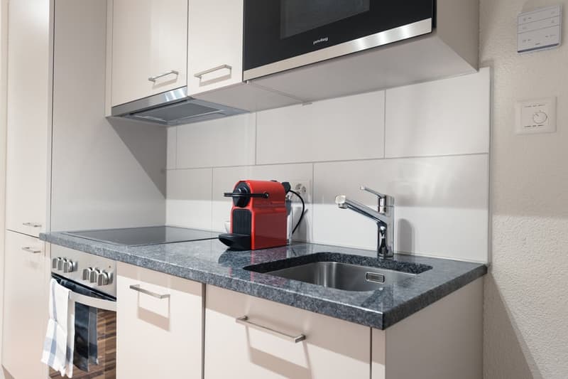 Modern kitchen with white cabinets, gray countertop, stainless steel sink, and a red coffee machine on the counter.