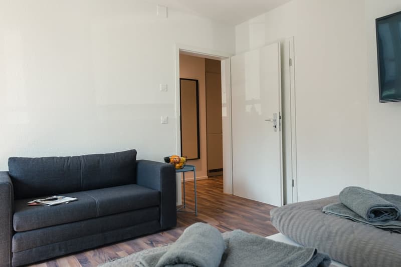Spacious living room with hardwood floors, a dark gray sofa, and a small side table with some fruit. The room has a door leading to what appears to be a balcony or terrace.