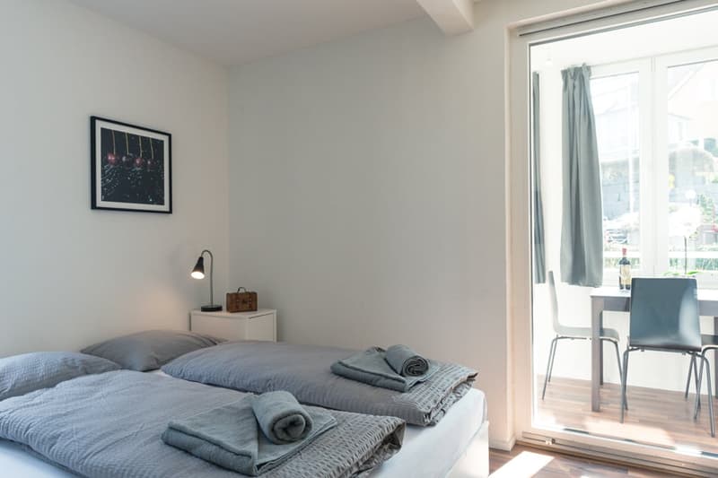 Spacious bedroom with large windows, grey bedding, nightstand with lamp, and a framed artwork on the wall.