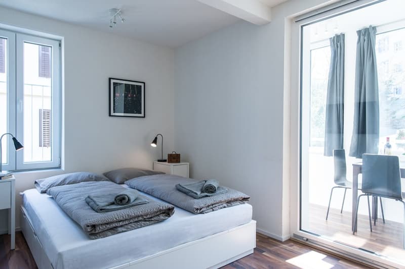 Spacious bedroom with large windows, grey bedding, side table, and artwork on the wall