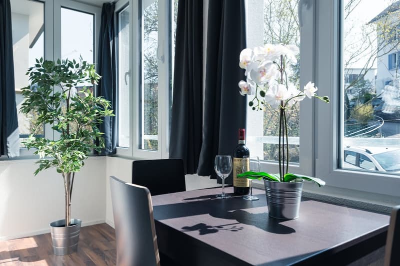 Spacious dining room with large windows overlooking a scenic outdoor view, black dining table and chairs, potted plant, orchid flower arrangement, and a bottle of wine with glasses on the table