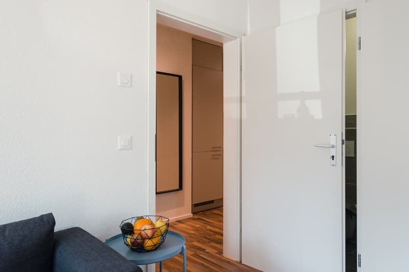 The image shows a modern and minimalist apartment interior. The room has white walls, hardwood floors, and a gray sofa. There is a small side table with a fruit bowl on it, and a doorway leading to another room or area of the apartment.