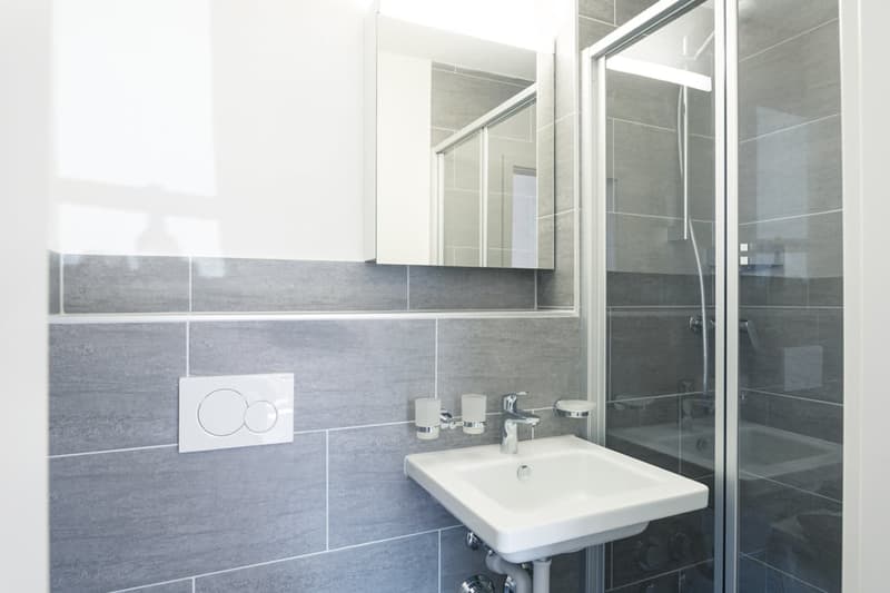 Modern bathroom with gray tile walls, white sink, and glass shower enclosure