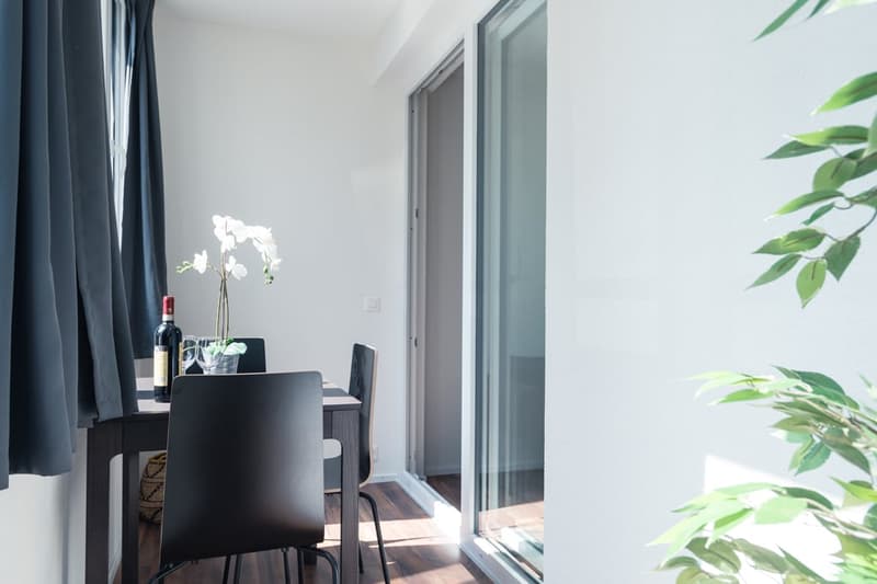 Modern dining area with a black table and chairs, a wine bottle and glasses, and a large window overlooking a balcony or terrace with greenery outside.