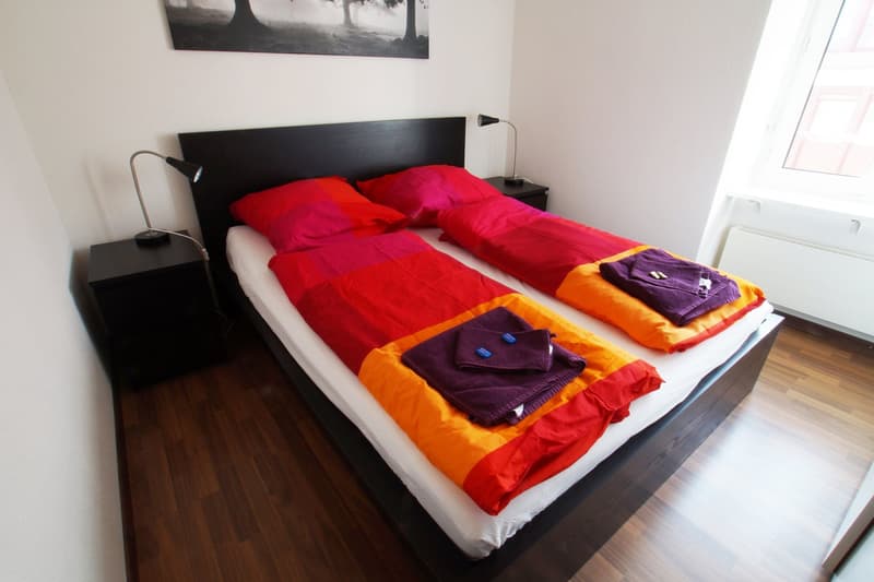 Spacious bedroom with a black wooden headboard, a large bed with colorful bedding, and two bedside tables with lamps. The room has hardwood flooring and a large window, suggesting the presence of a balcony or terrace.