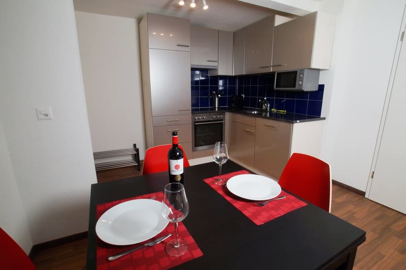 Modern kitchen with beige cabinets, black countertops, blue tile backsplash, oven, microwave, and dining table set with red placemats, plates, and wine glasses