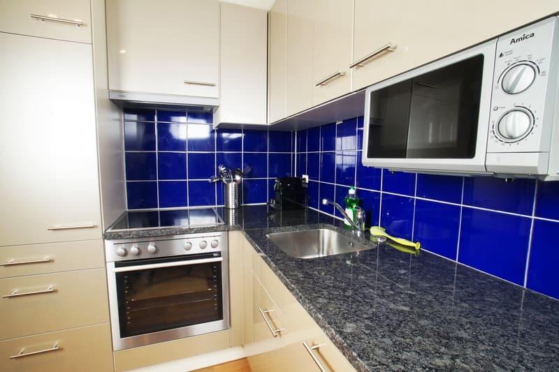 Modern kitchen with white cabinets, blue tile backsplash, granite countertops, stainless steel oven and microwave, and a sink with faucet