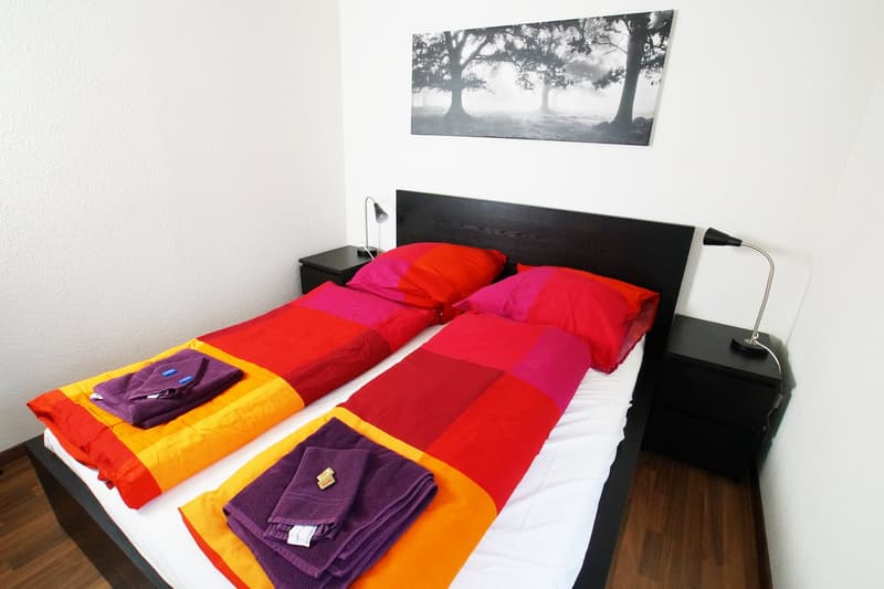 The image shows a modern bedroom with a black wooden headboard and a large colorful quilt on the bed. There is a black side table with a lamp, and a framed black and white landscape photograph on the wall. The room has a warm and cozy feel.