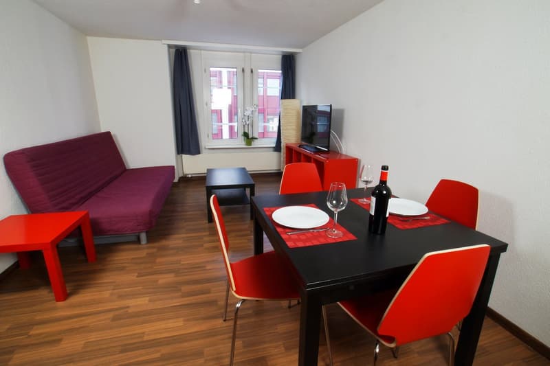 Spacious living room with hardwood floors, large windows, a purple sofa, a black coffee table, a TV stand, and a dining table with red chairs. The room also has a dishwasher and a bottle of wine on the table, indicating it is a combined living and dining